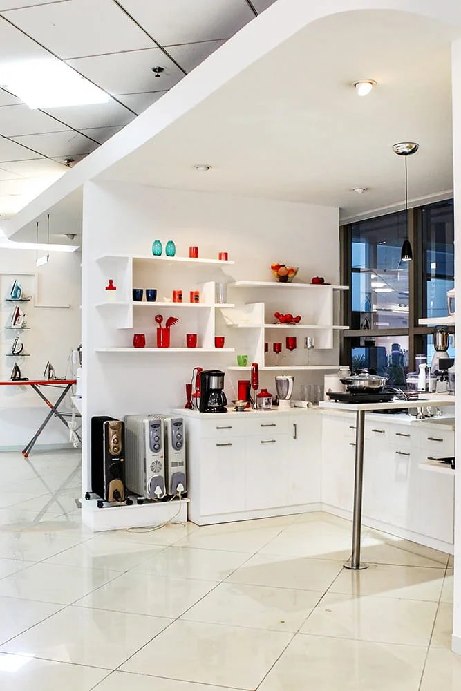 Vertical view of modular kitchen showcase featuring small domestic appliances, coffee maker, oil-filled room heaters, and custom white shelving photographed by Orange Images Photography for Havells India Limited Experiential Product Gallery in Noida, Uttar Pradesh, featuring small home appliances display, modern retail interior architecture, and interactive kitchen product exhibit.